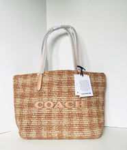 Load image into Gallery viewer, A Coach woven straw tote bag with light beige leather handles and a large metallic 'COACH' logo on the front, shown with its retail hangtag against a white background.